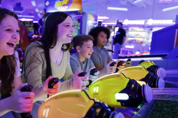 Group of four young players enjoying an arcade game with bright yellow controllers in a vibrant, illuminated gaming environment, reflecting the fun atmosphere of ClawCADE's arcade franchise experience.