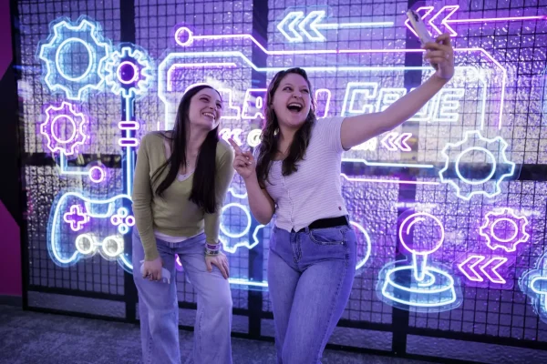 Two young women posing and taking a selfie in front of a vibrant neon gaming-themed backdrop featuring arcade elements, emphasizing the fun and social atmosphere of ClawCADE's modern arcade experience.